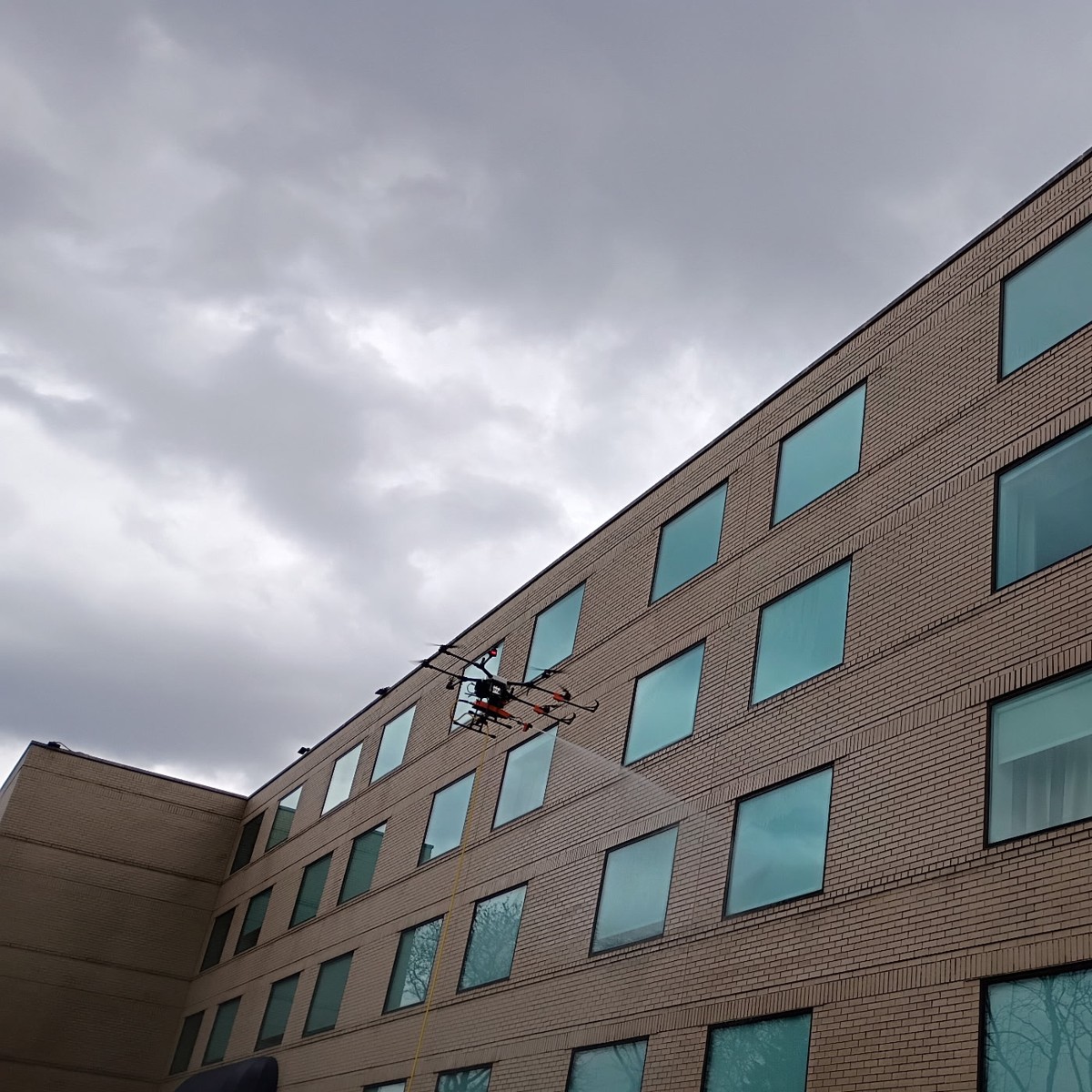 A tethered cleaning drone hovering beside the upper floors of a commercial building, spraying the brick and window glass