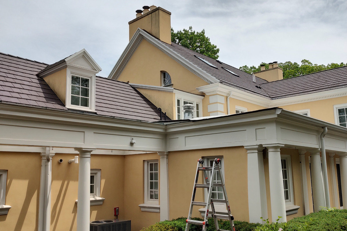 A ladder set against a large estate mid roof-wash, dark tile roof and tan stucco being cleaned