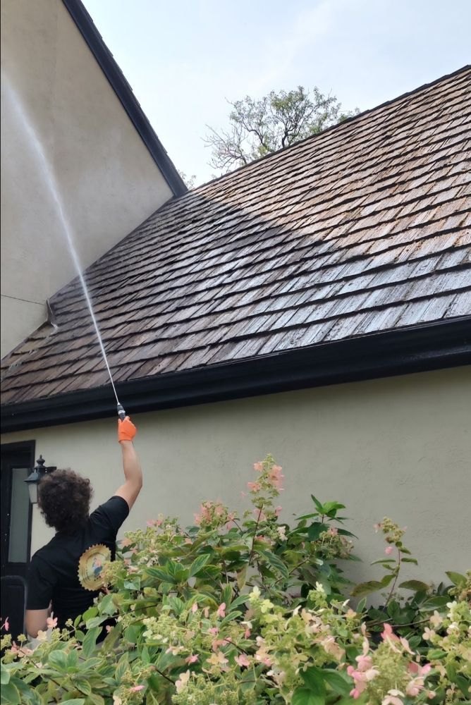 A crew member rinsing the roofline and dark gutters of a stucco home