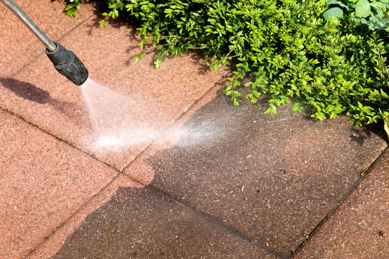 Close up of a pressure washer nozzle rinsing dirt off a red paver walkway beside a green hedge